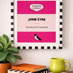 Pu&ograve; includere: Una stampa incorniciata di "Jane Eyre" di Charlotte Bront&euml;, con un design rosa e bianco e un bordo a scacchi bianco e nero. Una piccola illustrazione di un uccello &egrave; sulla copertina del libro. Una tazza verde, occhiali da sole e libri sono su una superficie di legno.