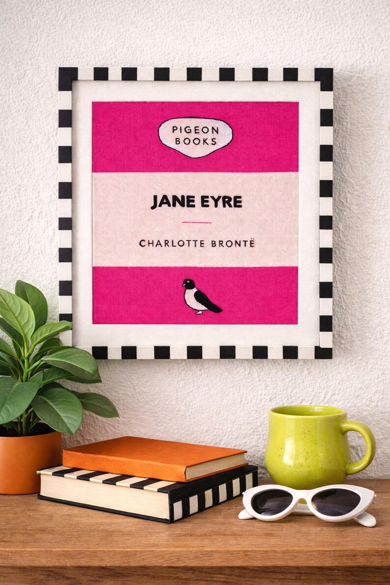 Pu&ograve; includere: Una stampa incorniciata di "Jane Eyre" di Charlotte Bront&euml;, con un design rosa e bianco e un bordo a scacchi bianco e nero. Una piccola illustrazione di un uccello &egrave; sulla copertina del libro. Una tazza verde, occhiali da sole e libri sono su una superficie di legno.