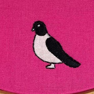 Pu&ograve; includere: Ricamo di uccello su tessuto rosa. L'uccello &egrave; nero e bianco, con cuciture dettagliate. Il tessuto &egrave; teso all'interno di un telaio da ricamo in legno, con un bordo bianco e rosa.