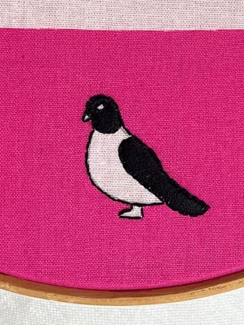 Pu&ograve; includere: Ricamo di uccello su tessuto rosa. L'uccello &egrave; nero e bianco, con cuciture dettagliate. Il tessuto &egrave; teso all'interno di un telaio da ricamo in legno, con un bordo bianco e rosa.