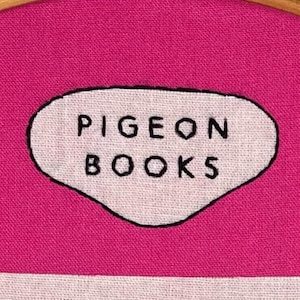 Pu&ograve; includere: Primo piano di un ricamo con la scritta "PIGEON BOOKS" in nero su un ovale bianco. L'ovale &egrave; posto su un tessuto rosa acceso, tenuto in un telaio da ricamo in legno.