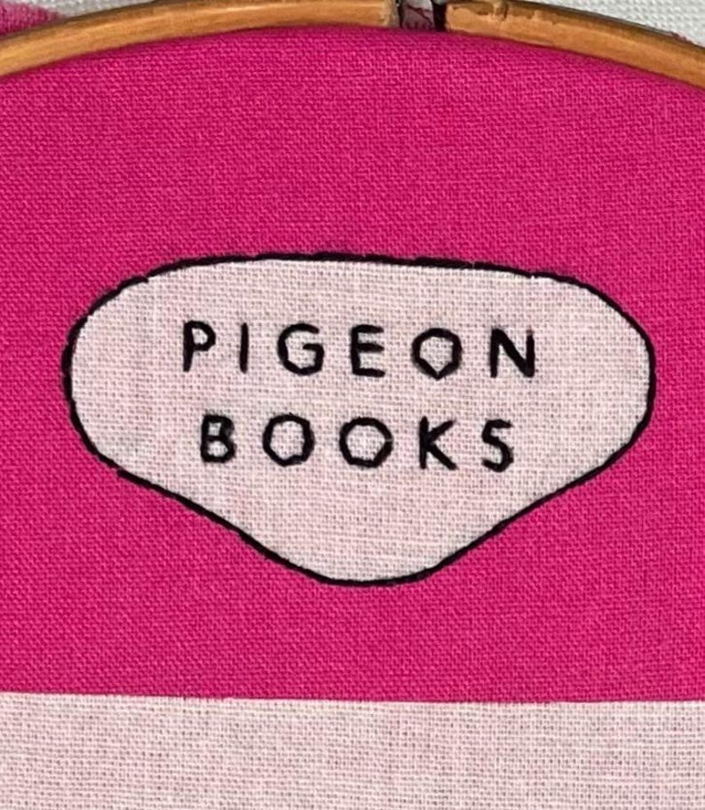 Pu&ograve; includere: Primo piano di un ricamo con la scritta "PIGEON BOOKS" in nero su un ovale bianco. L'ovale &egrave; posto su un tessuto rosa acceso, tenuto in un telaio da ricamo in legno.