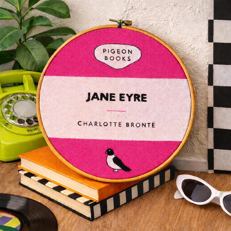 Pu&ograve; includere: Un'opera d'arte ricamata a cerchio rosa e bianca con il titolo del libro "Jane Eyre" di Charlotte Bront&euml;. Il design include un'illustrazione di un uccello e il testo "Pigeon Books". L'opera &egrave; esposta con libri, un telefono vintage e occhiali da sole.