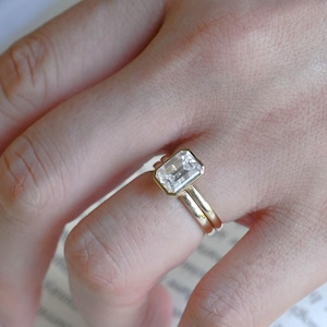 May include: A gold ring featuring a rectangular emerald-cut clear gemstone set in a bezel setting. The ring has a smooth, polished gold band and is worn on a finger, showcasing the clarity and faceted edges of the central stone.