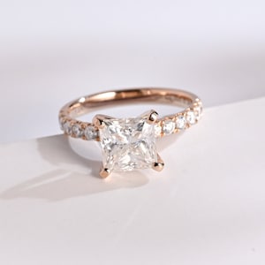 May include: A rose gold engagement ring with a square-shaped diamond center stone and a band of smaller diamonds.