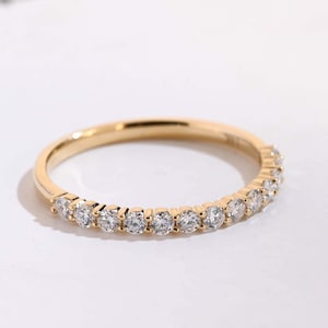 May include: A thin gold band ring featuring a row of small, round, clear faceted stones set along the top half of the band. The metal has a polished finish, and the stones are arranged in a continuous line, creating a sparkling, delicate appearance.