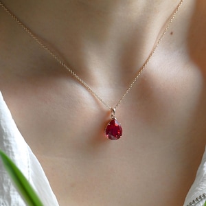 May include: A delicate gold chain necklace featuring a single, faceted, teardrop-shaped red gemstone pendant. The pendant is securely held in a simple metal setting, resting against the neck on a thin, polished gold-tone metal chain.