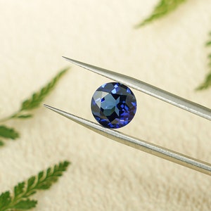 May include: A deep blue sapphire gemstone held by silver tweezers against a white background.