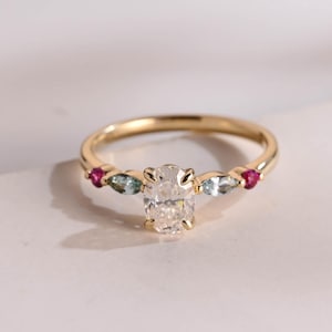 Op de afbeelding: Een gouden ring met een ovaalvormige diamant als middensteen, geflankeerd door twee kleine, lichtblauwe edelstenen en twee kleine, rode edelstenen. De ring heeft een delicate band en staat tegen een witte achtergrond.