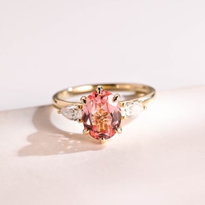May include: A gold ring featuring a pink and orange oval gemstone, flanked by two smaller pear-shaped gemstones.