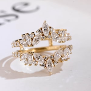 May include: A gold ring guard featuring two bands connected by a vertical bar. The design is adorned with marquise-cut and round-cut clear stones arranged in a V-shaped, leaf-like pattern along the top and bottom edges of the ring structure.