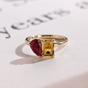 May include: A gold ring featuring a red heart-shaped gemstone and a yellow rectangular gemstone.