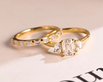 Op de afbeelding: Twee gouden ringen met diamanten. De grotere ring heeft een centrale smaragd geslepen diamant geflankeerd door twee kleinere ronde diamanten. De kleinere ring heeft een band van kleinere baguette geslepen diamanten.