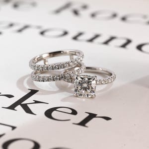 May include: Three diamond rings. One ring has a large, square-cut diamond. The other two are thin bands, each with small diamonds. The rings are on a white surface with blurred text in the background. The rings are made of silver.