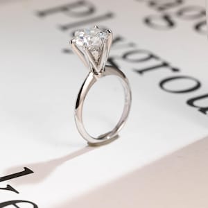 Puede incluir: Un anillo de compromiso plateado con un diamante grande y transparente. El diamante está engastado con seis garras, y la banda es lisa y pulida. El anillo se muestra sobre una superficie blanca con texto borroso en el fondo.