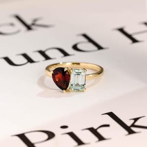 May include: A gold ring featuring a red pear-shaped gemstone and a green rectangular gemstone.
