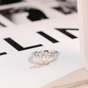 Puede incluir: Un anillo de compromiso de plata con un gran diamante central de talla esmeralda rectangular y una banda de diamantes más pequeños.