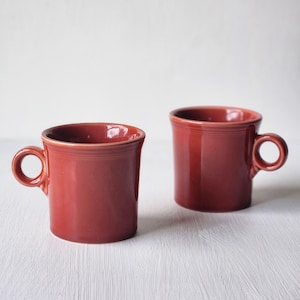 May include: Two red ceramic mugs with rounded handles. The mugs have a classic design with a slightly flared rim and a smooth, glossy finish. The mugs are displayed on a white surface, showcasing their vibrant color and simple elegance.
