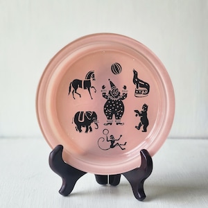 May include: A vintage pink plate with black circus-themed illustrations, including a horse, elephant, clown, seal, and monkey. The plate is displayed on a dark brown stand, showcasing a retro design.
