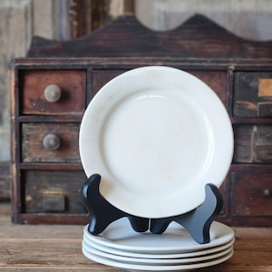 May include: A stack of six white ceramic plates with a textured surface. The plates are displayed on a black plate stand. The plates are in front of a wooden cabinet with drawers.