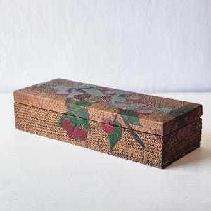 May include: A rectangular wooden box with a hinged lid. The box is brown with a woven texture and features a painted design of cherries and leaves in red, green, and purple. The box is set against a white background.