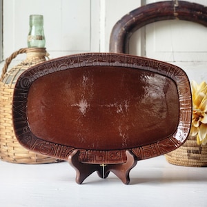 May include: An oval-shaped brown ceramic platter featuring a glossy finish and a wide, textured border decorated with repeating geometric and stylized face motifs. The platter is displayed upright on a small, dark brown wooden tripod stand.