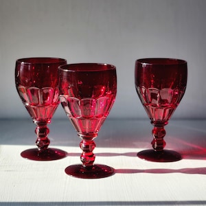 May include: Three red glass wine goblets with a faceted design. The goblets are on a white surface.