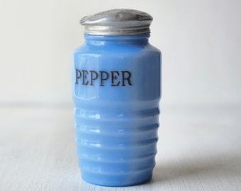 May include: A light blue glass pepper shaker with a silver metal lid. The word "PEPPER" is printed in black on the front. The shaker has a ribbed design around the body.