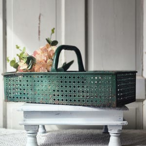 May include: A vintage green metal basket with a handle, filled with faux flowers, sits on a small, white, distressed wooden table. The basket has a perforated design. The table is on a white rug.
