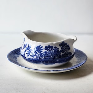 May include: A white ceramic gravy boat with a blue Willow pattern, resting on a matching saucer. The boat has a spout and handle, and the set features a classic blue and white landscape design.