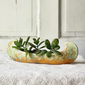 May include: A canoe-shaped ceramic planter featuring a mottled glaze in shades of pale green and warm orange. The vessel contains a small green succulent plant with thick, rounded leaves, positioned centrally within the elongated, curved structure.