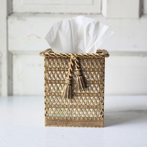 May include: A rectangular tissue box cover featuring a metallic gold finish with an intricate lattice pattern. The top edge is bordered by a twisted rope design, and two decorative gold tassels hang from the front center. White tissues protrude.