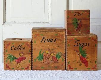May include: A set of four square wooden canisters with lids, featuring hand-painted rooster illustrations and black lettering. The containers are labeled Coffee, Flour, Tea, and Sugar, each decorated with red and green floral accents along the edges.