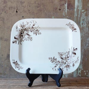 May include: A rectangular, off-white serving platter with rounded corners, adorned with brown floral patterns in each corner. The platter is displayed on a black stand, showcasing its vintage design. The background is a rustic wooden surface.