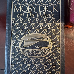 Może przedstawiać: Twarda oprawa książki "Moby Dick or The Whale" Hermana Melville'a. Okładka jest ciemnoniebieska ze złotymi literami i ilustracją wieloryba. Wz&oacute;r zawiera wzory fal.