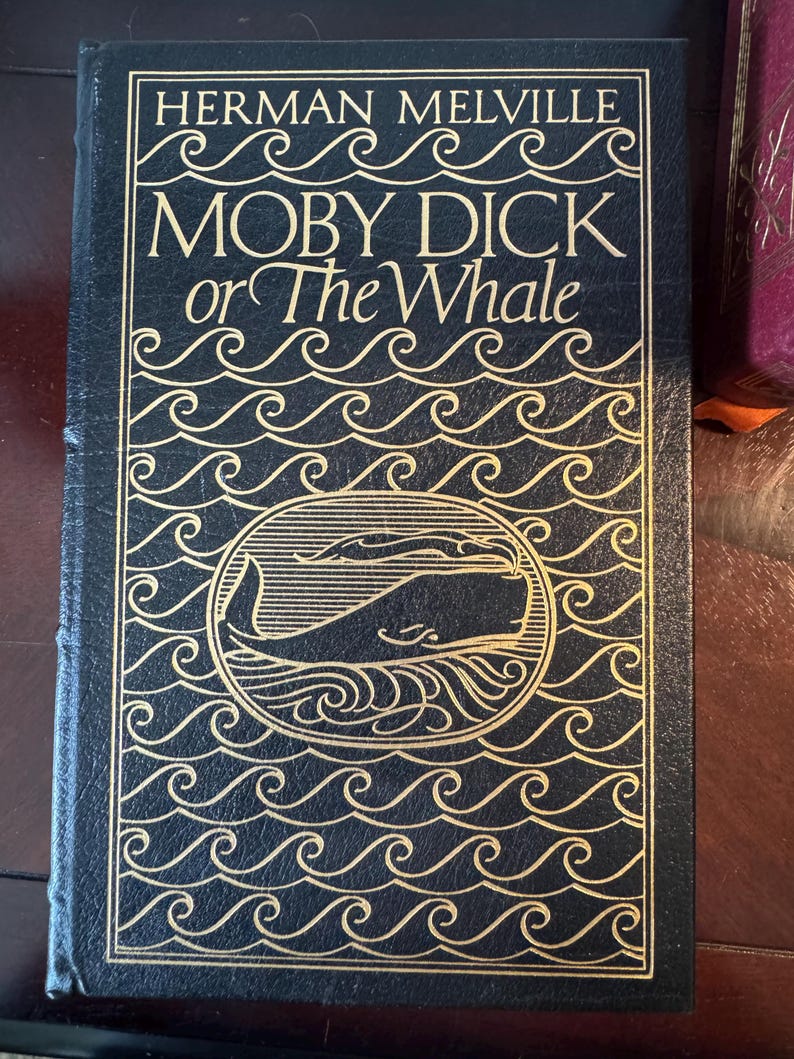 Może przedstawiać: Twarda oprawa książki "Moby Dick or The Whale" Hermana Melville'a. Okładka jest ciemnoniebieska ze złotymi literami i ilustracją wieloryba. Wz&oacute;r zawiera wzory fal.