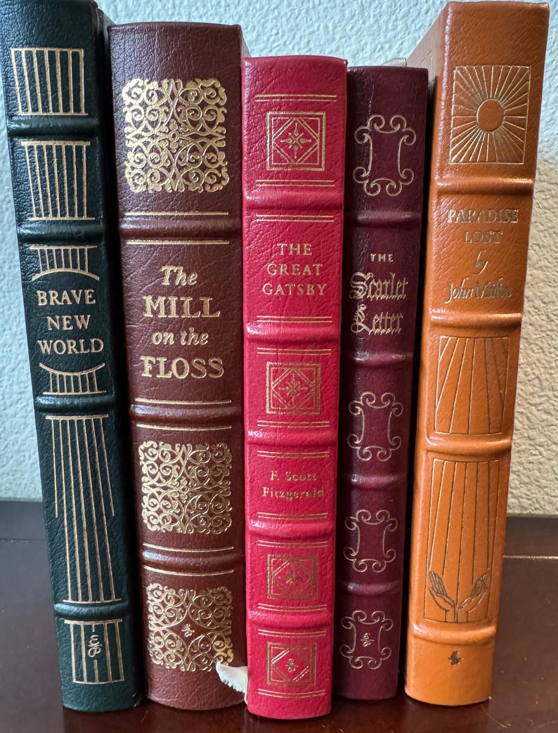 Może przedstawiać: Kolekcja klasycznych książek ze złotymi literami i zdobieniami. Tytuły to m.in. "Brave New World", "The Mill on the Floss", "The Great Gatsby", "The Scarlet Letter" i "Paradise Lost". Książki mają okładki w r&oacute;żnych kolorach.