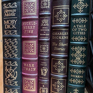 Może przedstawiać: Kolekcja klasycznych książek z pozłacanymi tytułami i zdobieniami. Widoczne tytuły to "Moby Dick", "Huckleberry Finn", "Alicja w Krainie Czar&oacute;w", "The Short Stories" i "Opowieść o dw&oacute;ch miastach". Grzbiety są w kolorach bordowym, brązowym i ciemnozielonym.