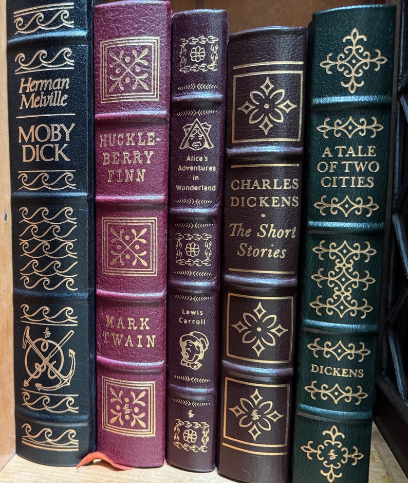 Może przedstawiać: Kolekcja klasycznych książek z pozłacanymi tytułami i zdobieniami. Widoczne tytuły to "Moby Dick", "Huckleberry Finn", "Alicja w Krainie Czar&oacute;w", "The Short Stories" i "Opowieść o dw&oacute;ch miastach". Grzbiety są w kolorach bordowym, brązowym i ciemnozielonym.