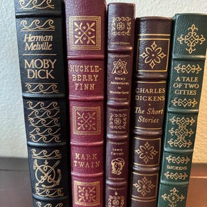 Może przedstawiać: Kolekcja pięciu klasycznych książek z dekoracyjnymi okładkami. Tytuły to "Moby Dick", "Huckleberry Finn", "Alicja w Krainie Czar&oacute;w", "The Short Stories" i "A Tale of Two Cities". Książki mają złote litery i ozdobne wzory.