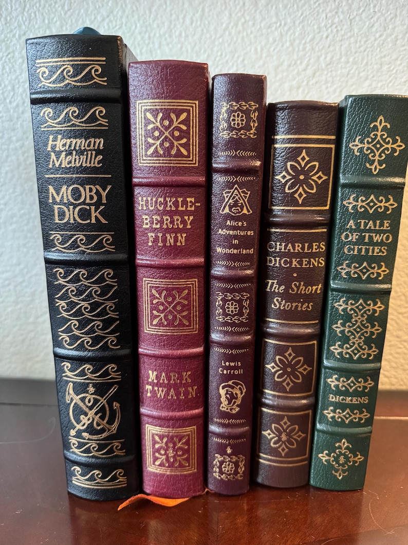 Może przedstawiać: Kolekcja pięciu klasycznych książek z dekoracyjnymi okładkami. Tytuły to "Moby Dick", "Huckleberry Finn", "Alicja w Krainie Czar&oacute;w", "The Short Stories" i "A Tale of Two Cities". Książki mają złote litery i ozdobne wzory.