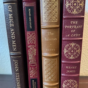 Może przedstawiać: Cztery książki w twardej oprawie ze złotymi literami i dekoracyjnymi wzorami. Tytuły to "Of Mice and Men", "Romeo and Juliet", "The Talisman" i "The Portrait of a Lady". Książki mają sk&oacute;rzane okładki w odcieniach brązu i bordo.