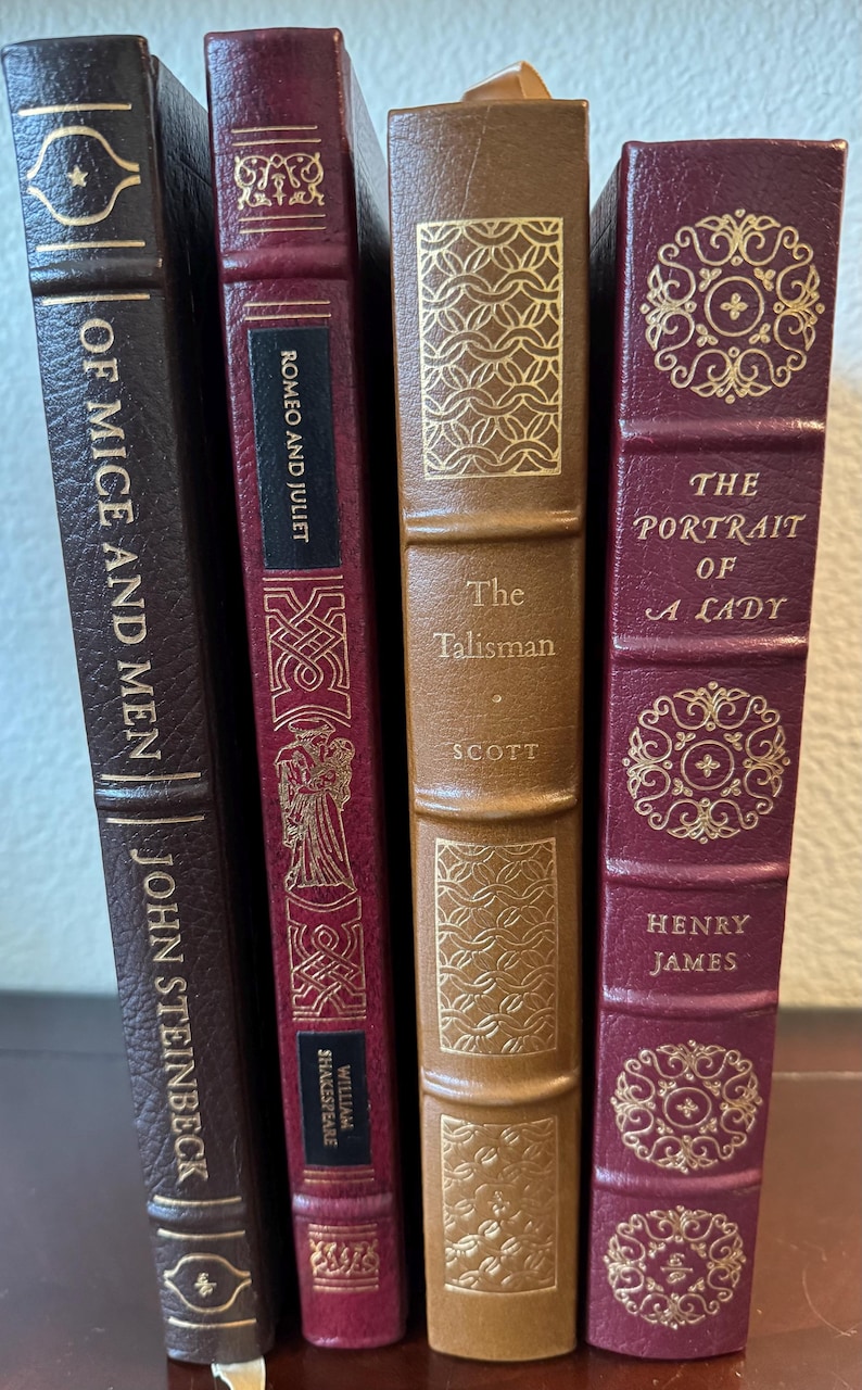 Może przedstawiać: Cztery książki w twardej oprawie ze złotymi literami i dekoracyjnymi wzorami. Tytuły to "Of Mice and Men", "Romeo and Juliet", "The Talisman" i "The Portrait of a Lady". Książki mają sk&oacute;rzane okładki w odcieniach brązu i bordo.