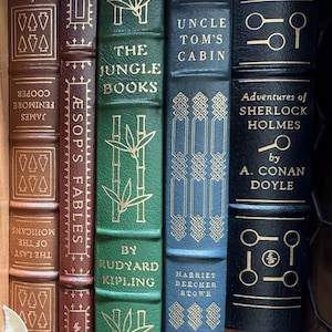 Może przedstawiać: Biały ceramiczny kubek z czarnym uchwytem i napisem "But First, Coffee" czarną, pogrubioną czcionką. Kubek stoi na drewnianej powierzchni. Kubek ma około 10 cm wysokości.