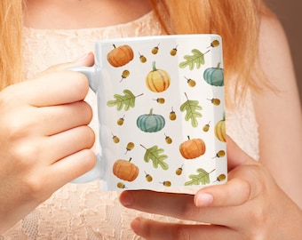 Puede incluir: Una taza de cerámica blanca con un patrón repetitivo de calabazas naranjas y verde azulado, hojas de roble verdes y pequeñas bellotas marrones. La taza tiene un acabado liso y un asa curva en el lateral con un diseño otoñal.