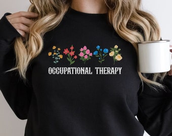 Könnte beinhalten: Schwarzer Pullover mit einer Reihe bunter Blumen und dem Aufdruck "OCCUPATIONAL THERAPY" auf der Vorderseite.