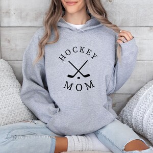 May include: Gray hooded sweatshirt with the text "HOCKEY MOM" in black lettering and a graphic of two crossed hockey sticks and a puck.