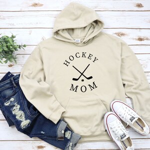 May include: A cream-colored hoodie with a black graphic that says "HOCKEY MOM" with two crossed hockey sticks and a puck.