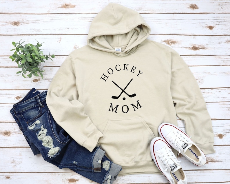 May include: A cream-colored hoodie with a black graphic that says "HOCKEY MOM" with two crossed hockey sticks and a puck.