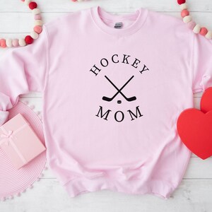 May include: A light pink sweatshirt with the words "HOCKEY MOM" in black, along with crossed hockey sticks and a puck. The sweatshirt is a crew neck style.
